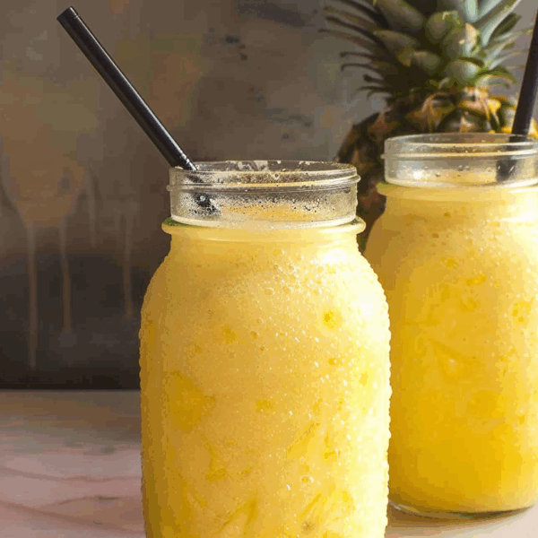 Easy Coconut Pineapple Smoothie With Coconut Milk: Irresistibly Delicious 7 Two mason jars filled with bright yellow pineapple coconut smoothie, topped with black straws, sitting on a marble counter with a whole pineapple in the background.